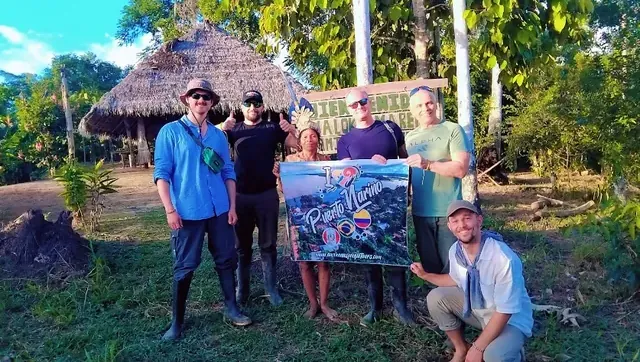 Grupo de visitantes con un cartel de Puerto Nariño frente a una maloca en la comunidad tikuna de Paraíso
